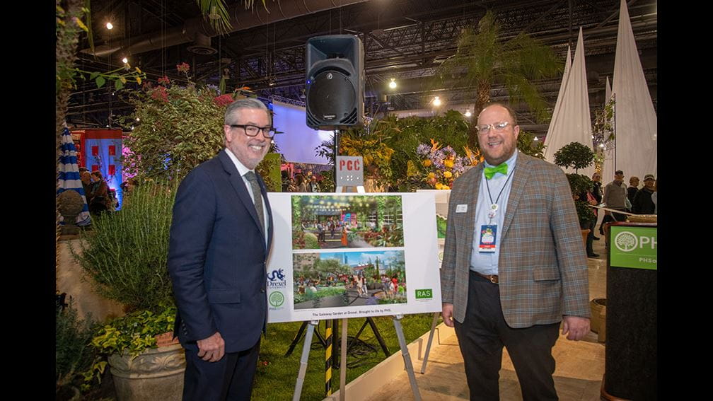 President John Fry posing next to Gateway Garden announcement poster board at Philadelphia Flower Show. He is accompanied by a someone wearing a lanyard.