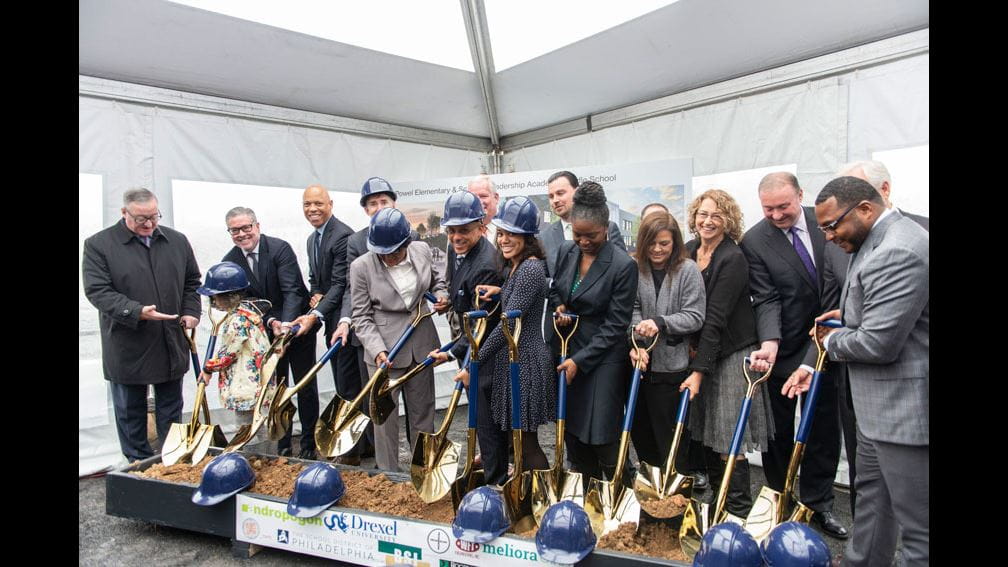 President John Fry posing at the K8 School Groundbreaking event with former Philly mayor John Kennedy, other city officials, and young child under white event tent. All are posing with shovels and some are wearing hard hats.