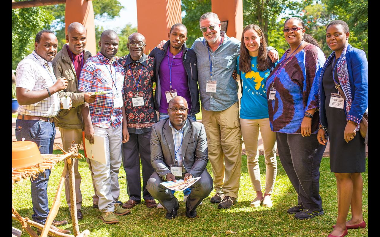 President John Fry standing with members of World Vision during Southern Africa Tour. They are outside in front of what appears to be a small courtyard with trees in the background.