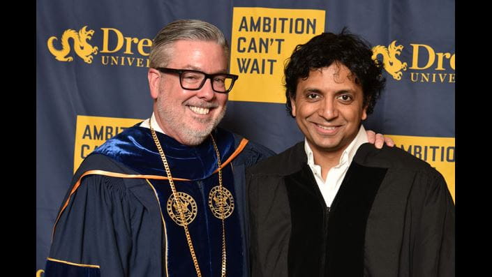 President John Fry dressed in commencement regalia standing next to film director M. Night Shyamalan. Both are standing in front of Drexel’s Ambition Can’t Wait backdrop.