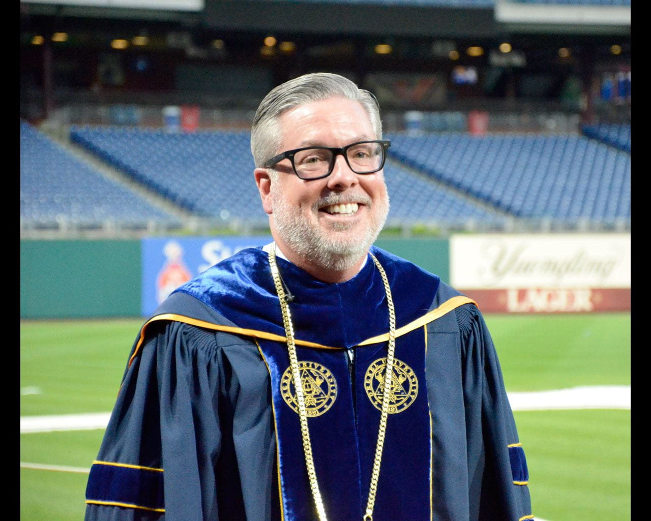 Portrait of John Fry standing in Citizens Bank Park while wearing commencement regalia.