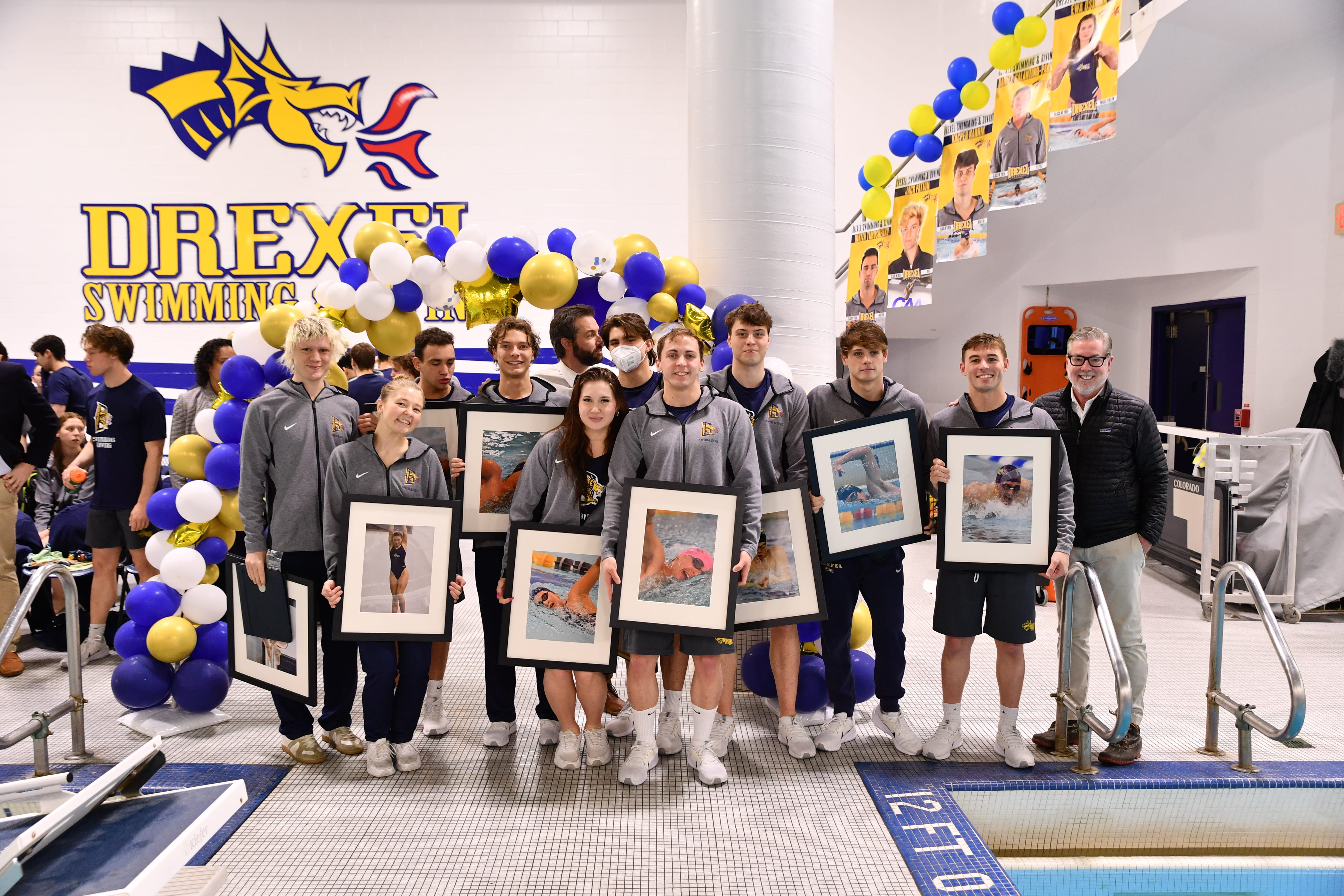 President John Fry standing next to members of Swimming & Diving team in gymnasium. Each team member is holding a framed picture of themselves competing.