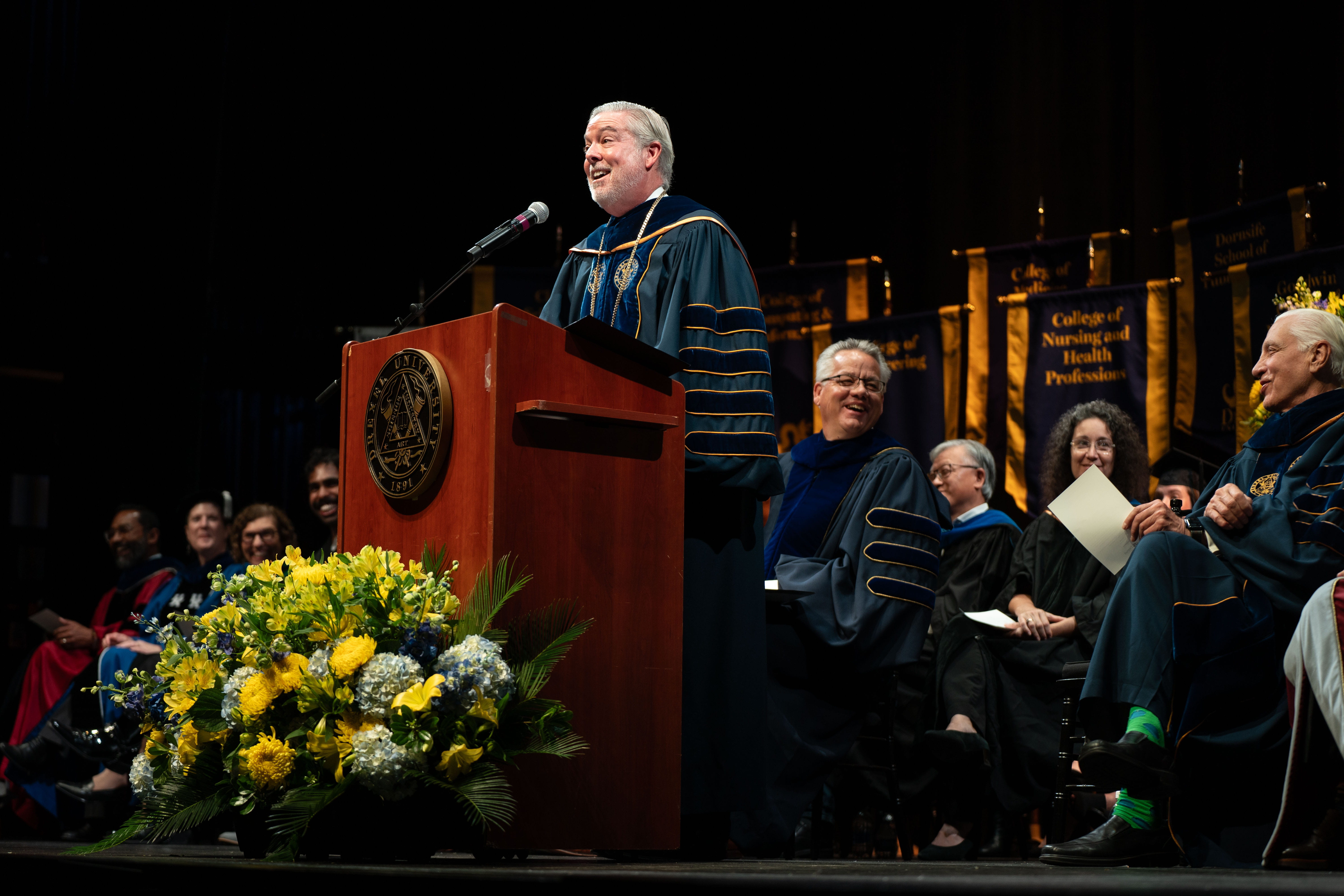 President John Fry giving speech at Convocation. Sitting behind him are various members of the Office of the Provost, including Paul E. Jensen, PhD.