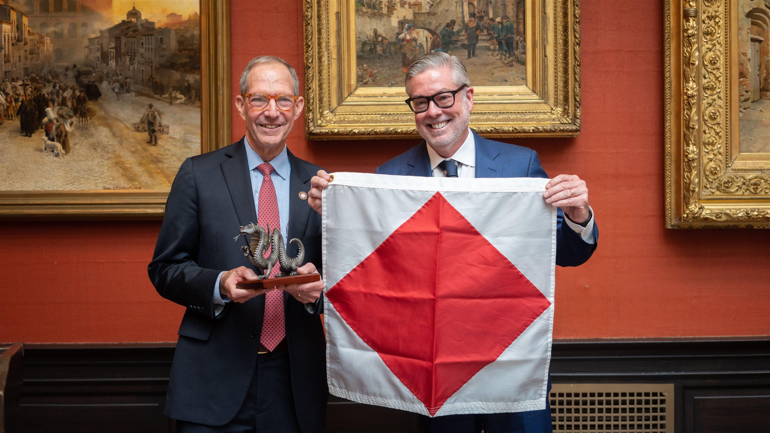 Salus University President Michael Mittelman (left) and Drexel University President John Fry standing next to each other in A.J. Drexel Picture Gallery. Michael is holding Drexel’s Mario the dragon  figurine while John holds Salus’ flag (white background with red diamond in the center).