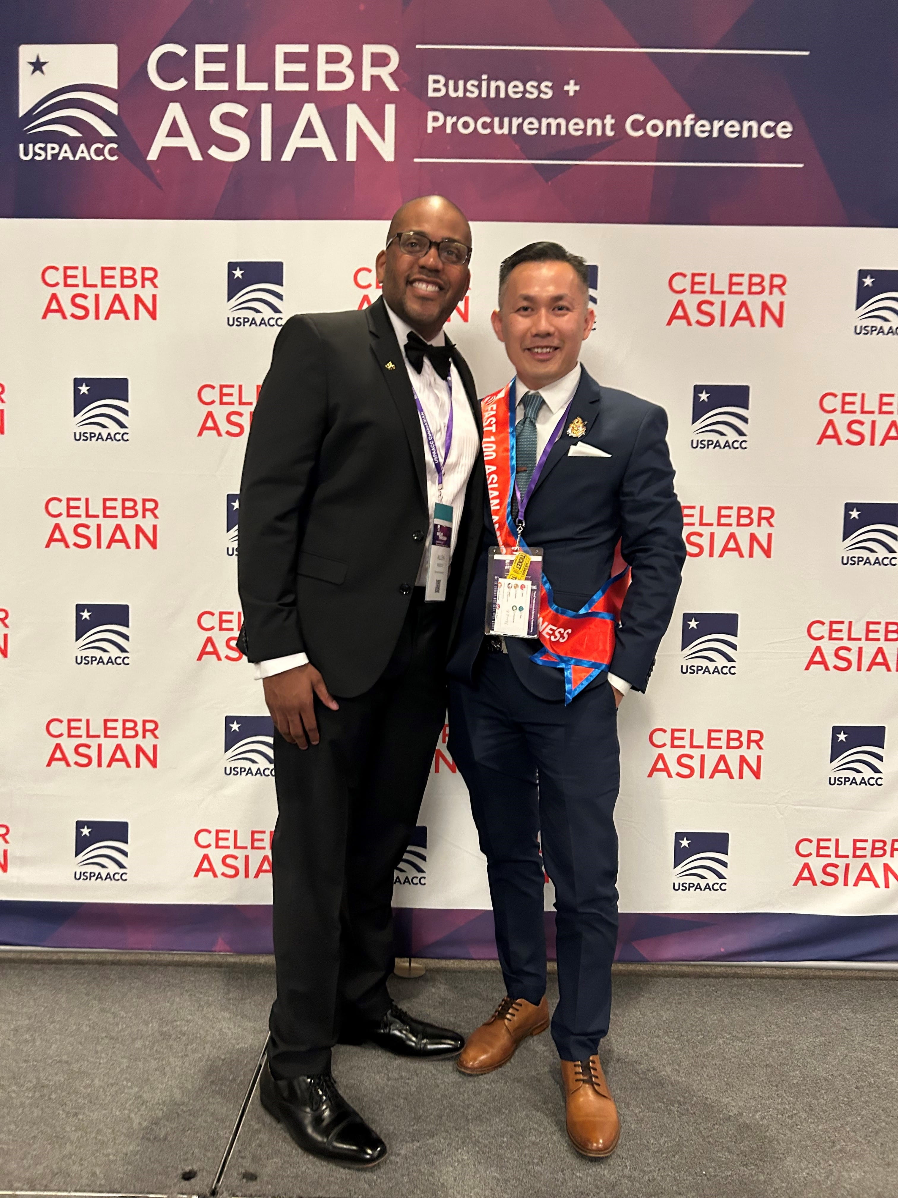 A Black man in a suit stands next to an Asian man in a suit with a red sash in front of a ppanel that reads "CelebrAsian."