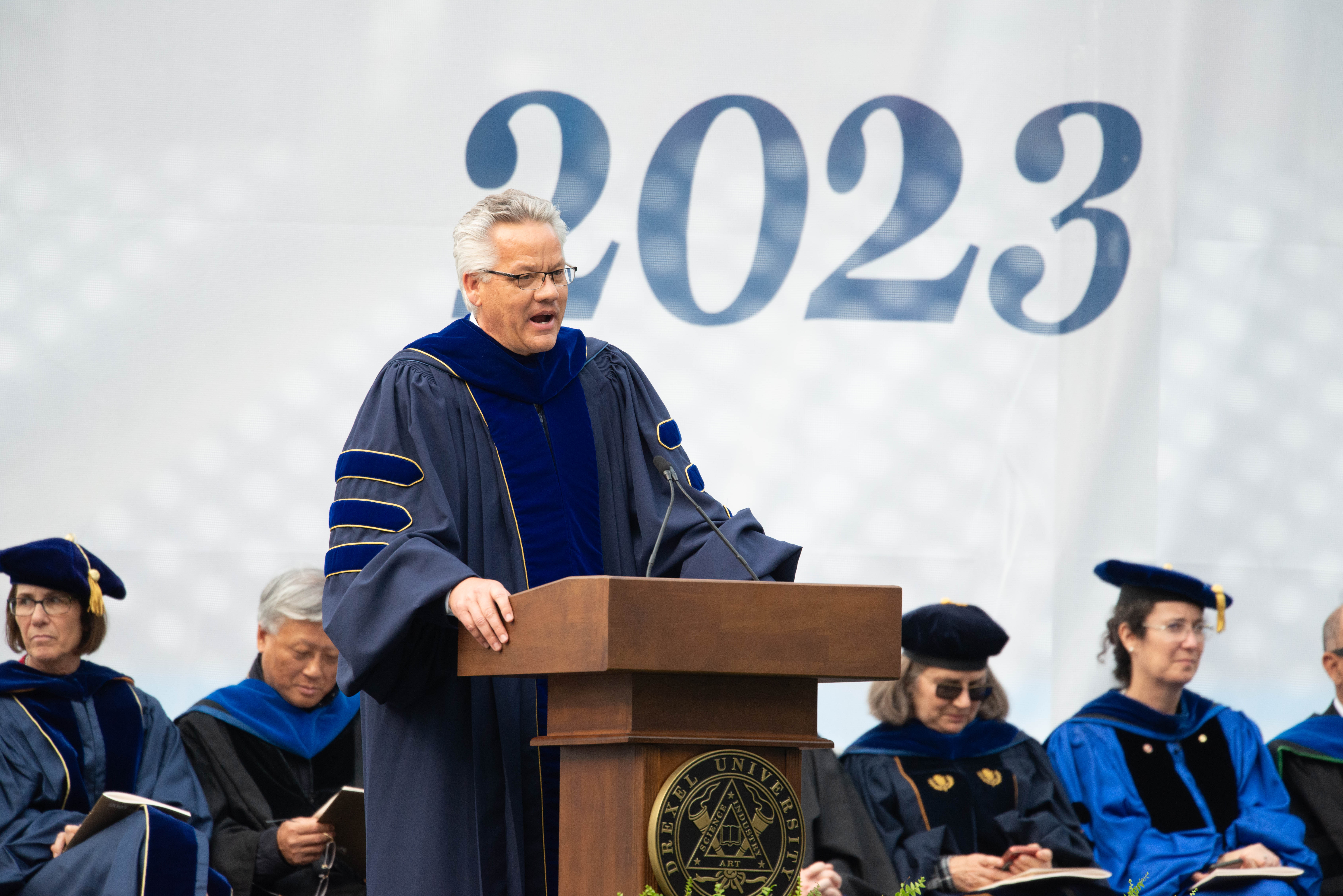 Provost Paul Jensen addresses graduates in front of people with caps and gowns and 2023 on the white background.