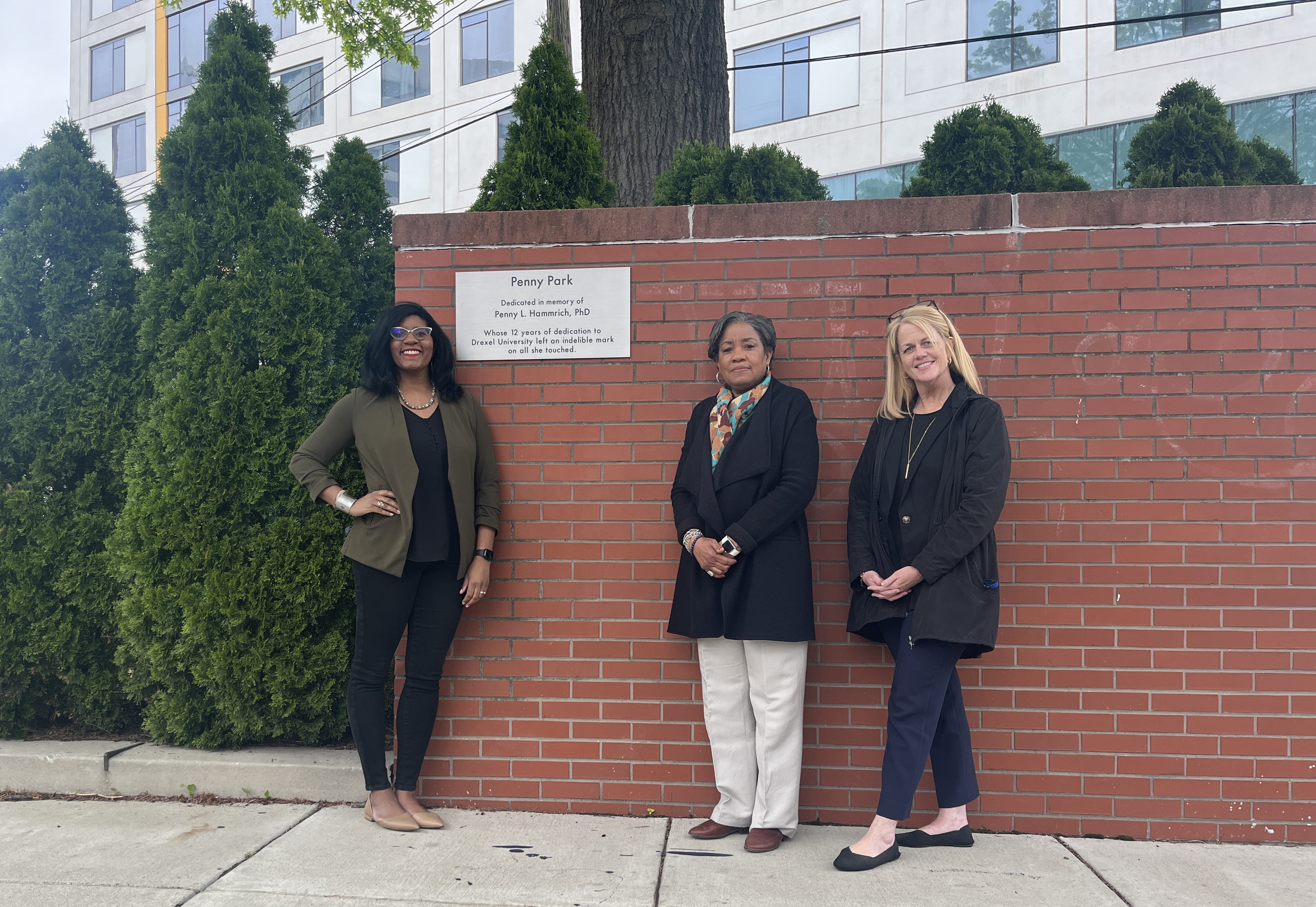 Three women stand in front of a brick wall with a plaque, next to trees.