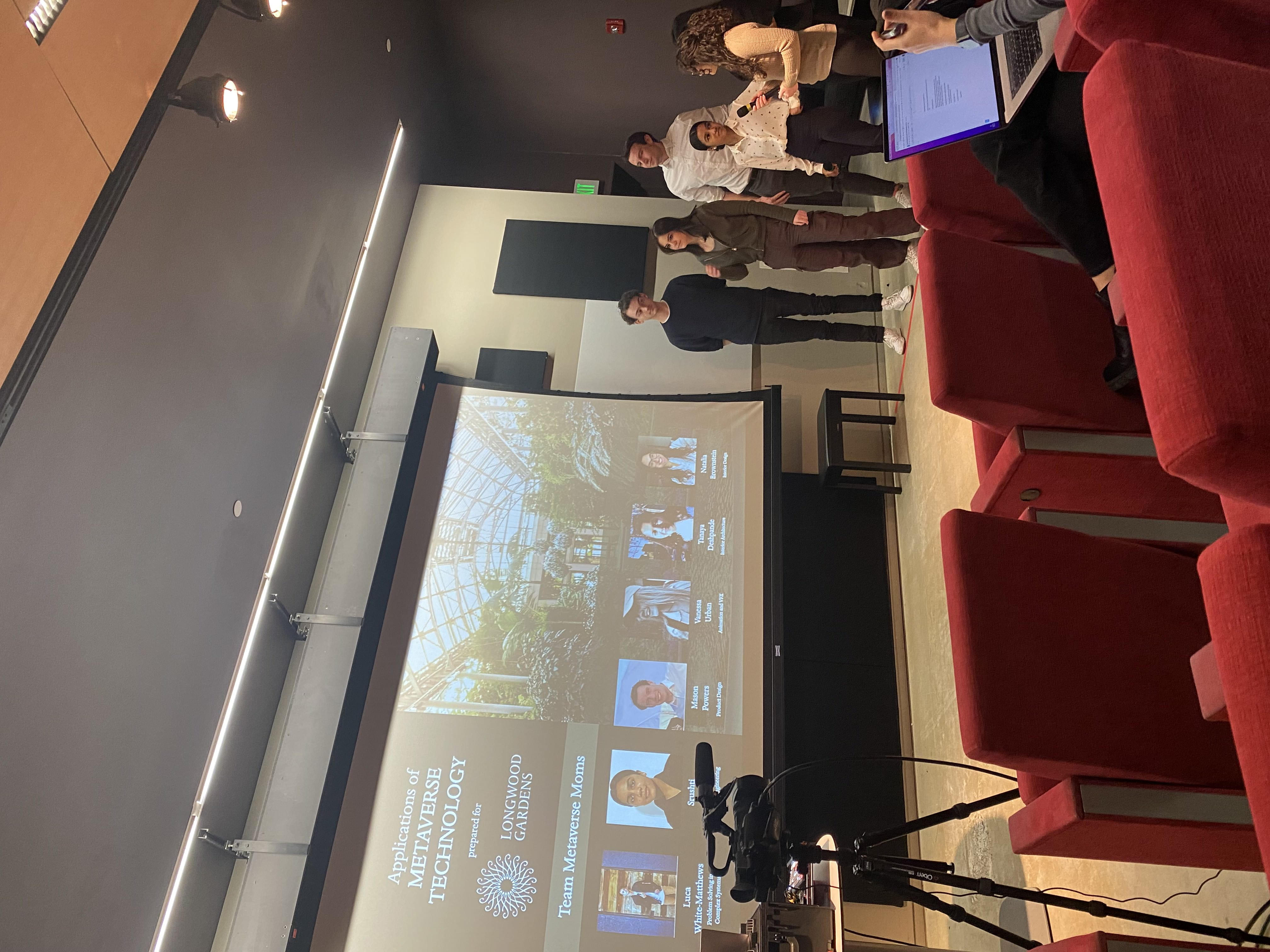 A team of students presenting in front of a projection screen that reads "Applications of Metaverse Technology"