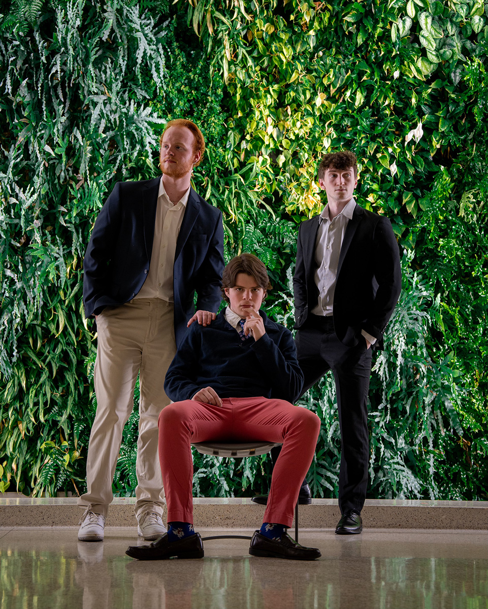 Three male students dressed in business attire pose in front of a wall of plants.