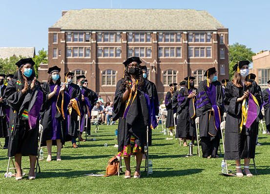 Three columns of graduates clapping their hands on a green field with a brick academic building in the background.