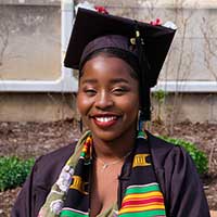 Photo of Kudzai, a Black woman smiling at the camera. She is wearing graduation regalia including a kente cloth. 