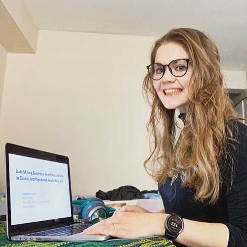 PhD student Elizabeth Campbell is photographed working from her home. She is seated with a laptop on her table, wearing glasses and smiling at the camera. 