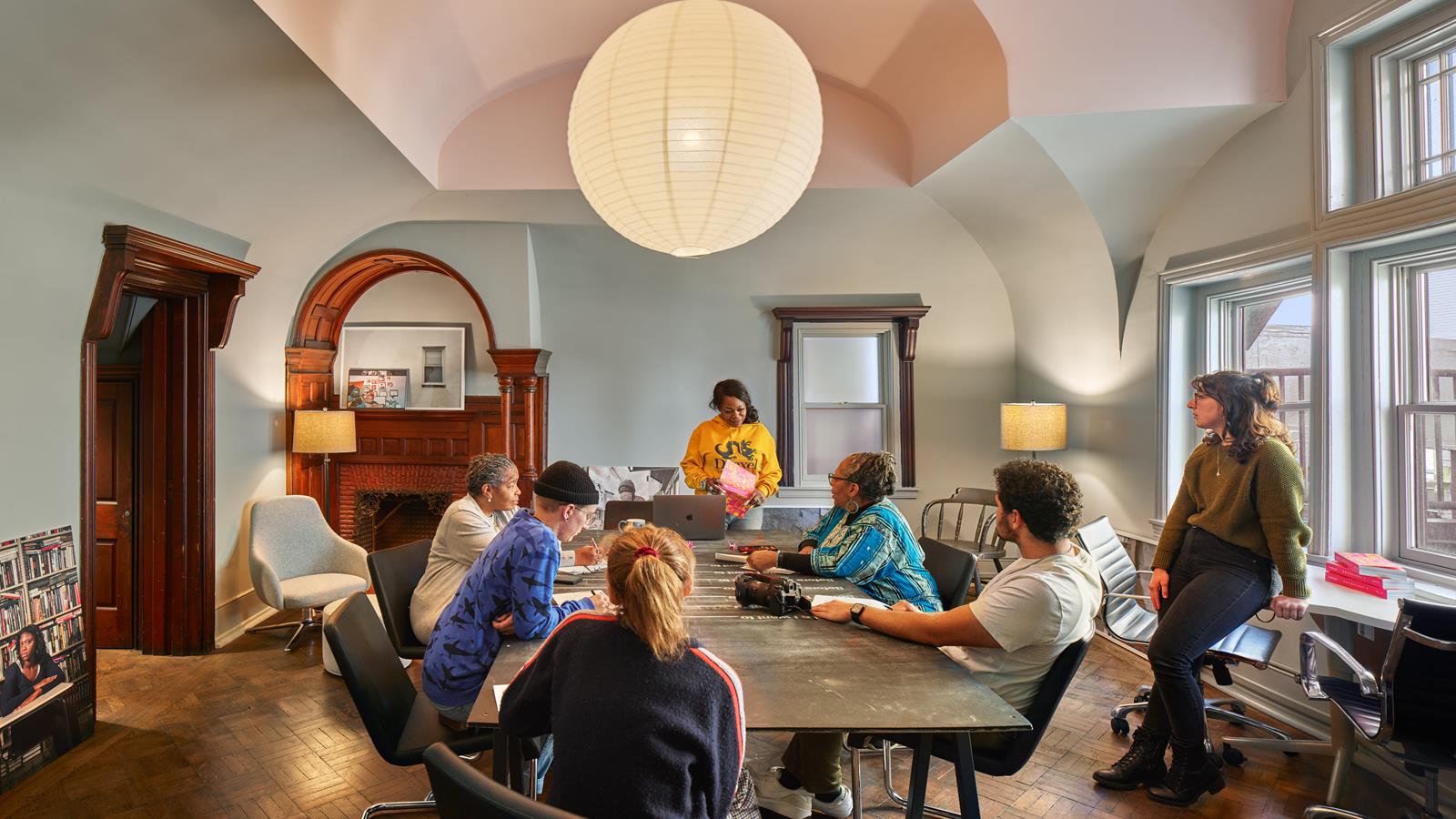 The Writers Room in Ross Commons. a cozy room with wooden furniture, a paper lantern light, and arched ceilings. Students sit around a table and discuss their work.