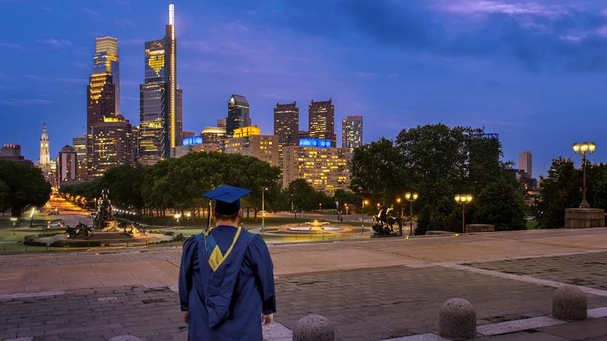 Drexel University graduate in cap and gown looking at Philadelphia skyline at dusk