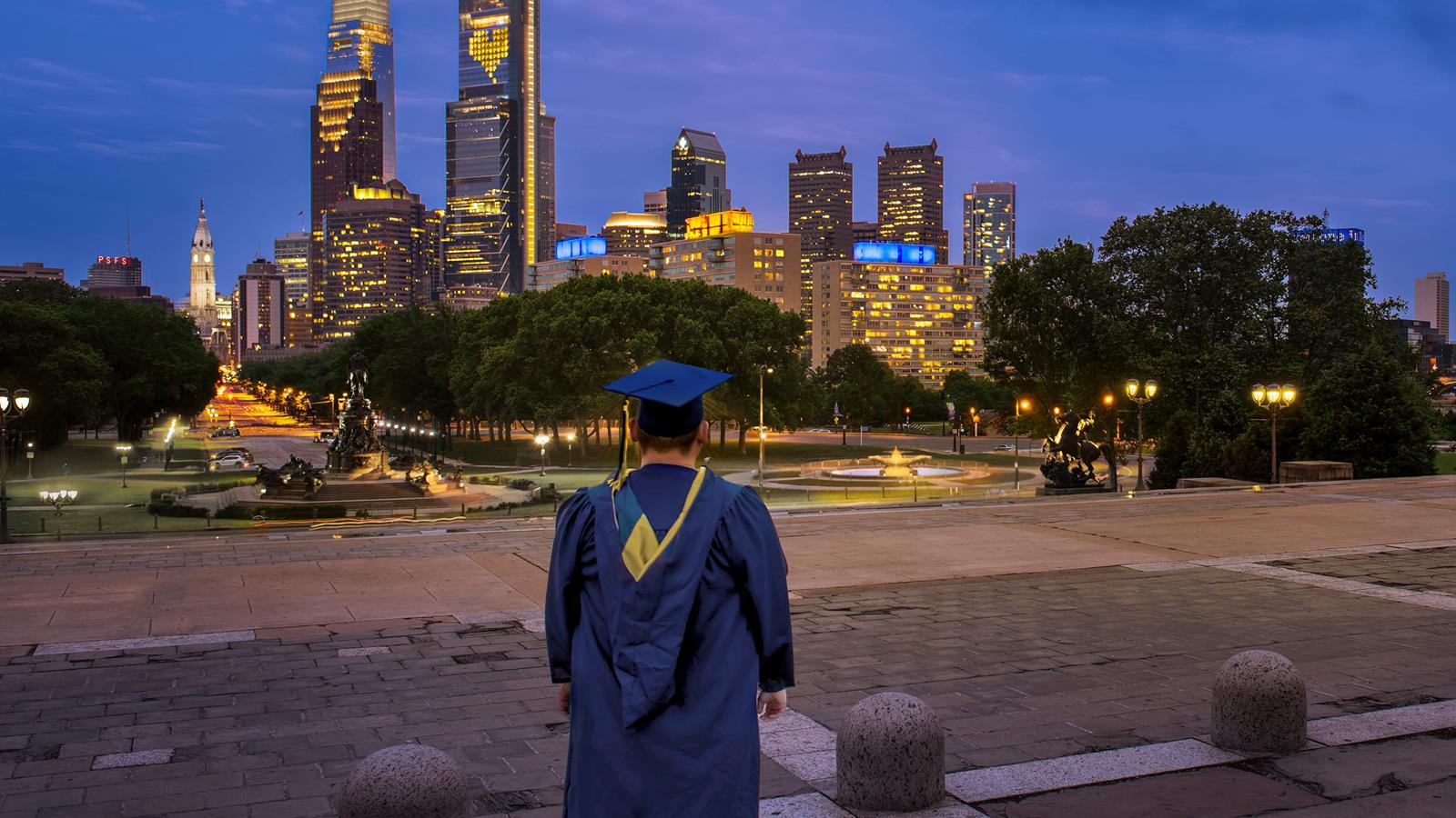 Drexel University graduate in cap and gown looking at Philadelphia skyline at dusk