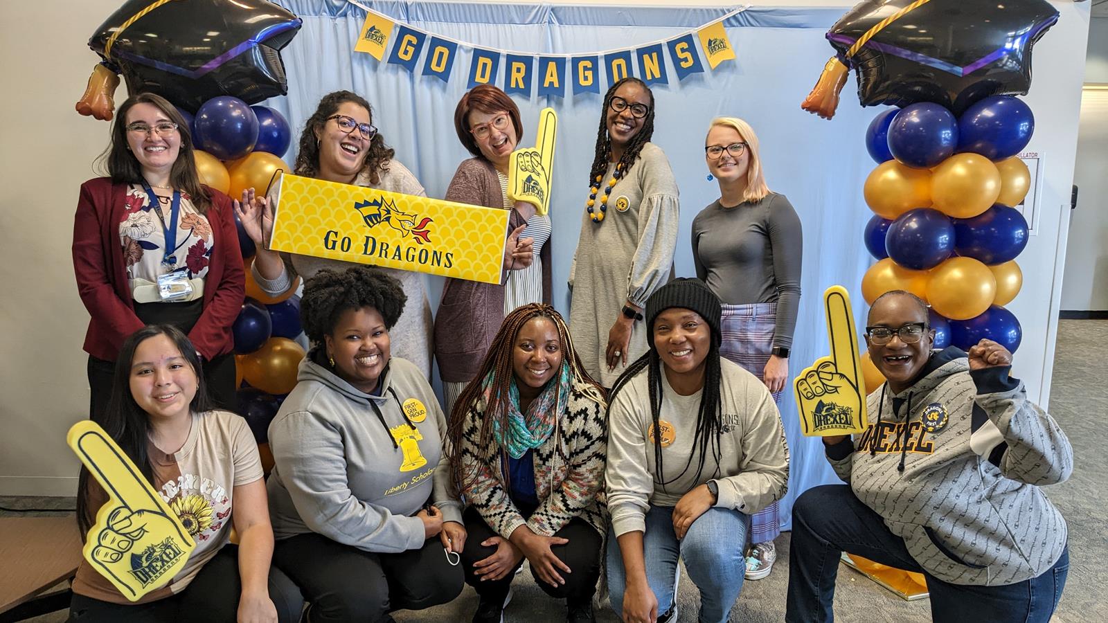 10 students, faculty, and staff pose in front of a blue backdrop with Drexel gear and Go Dragons! signs with balloons flanking the group
