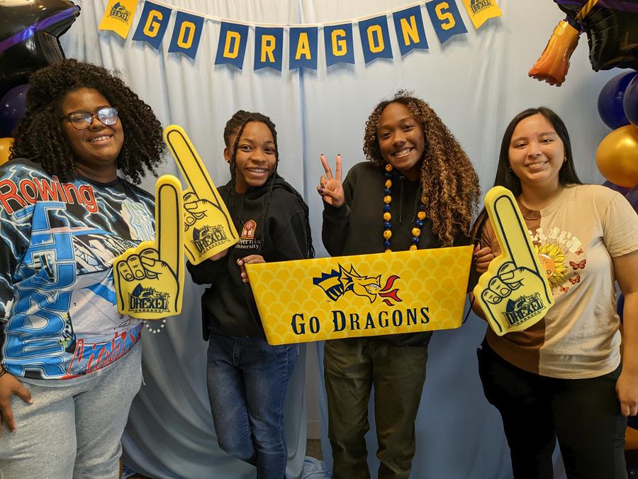 4 students pose in front of a blue backdrop with Drexel gear and Go Dragons! signs