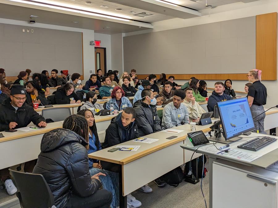 One student stands at the front of a classroom, speaking to 50 other students seated at long rows of desks