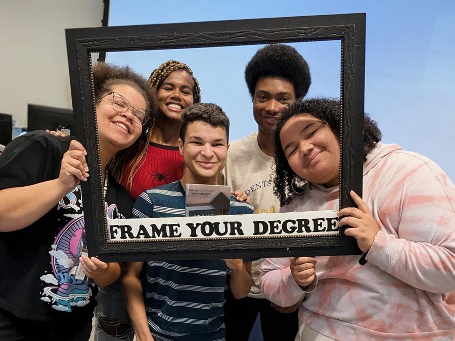 Five students pose, their faces framed in an oversized picture frame that reads "Frame Your Degree" across the bottom.
