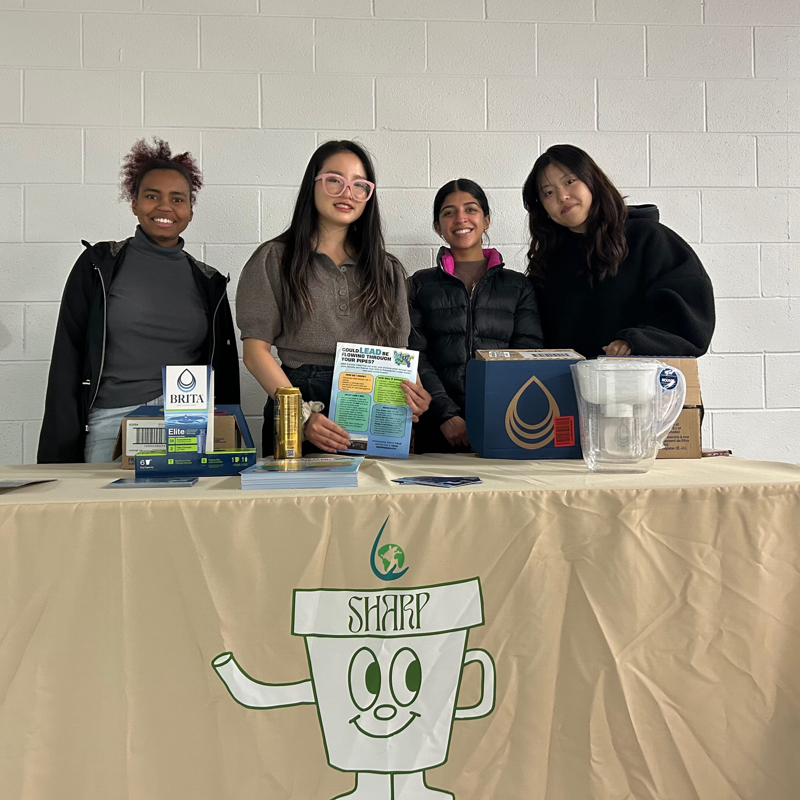 A group of four students pose behind a table. They have diverse skin and hair colors and styles and are smiling. The table is covered by a tan tablecloth with a white logo for SHARP.