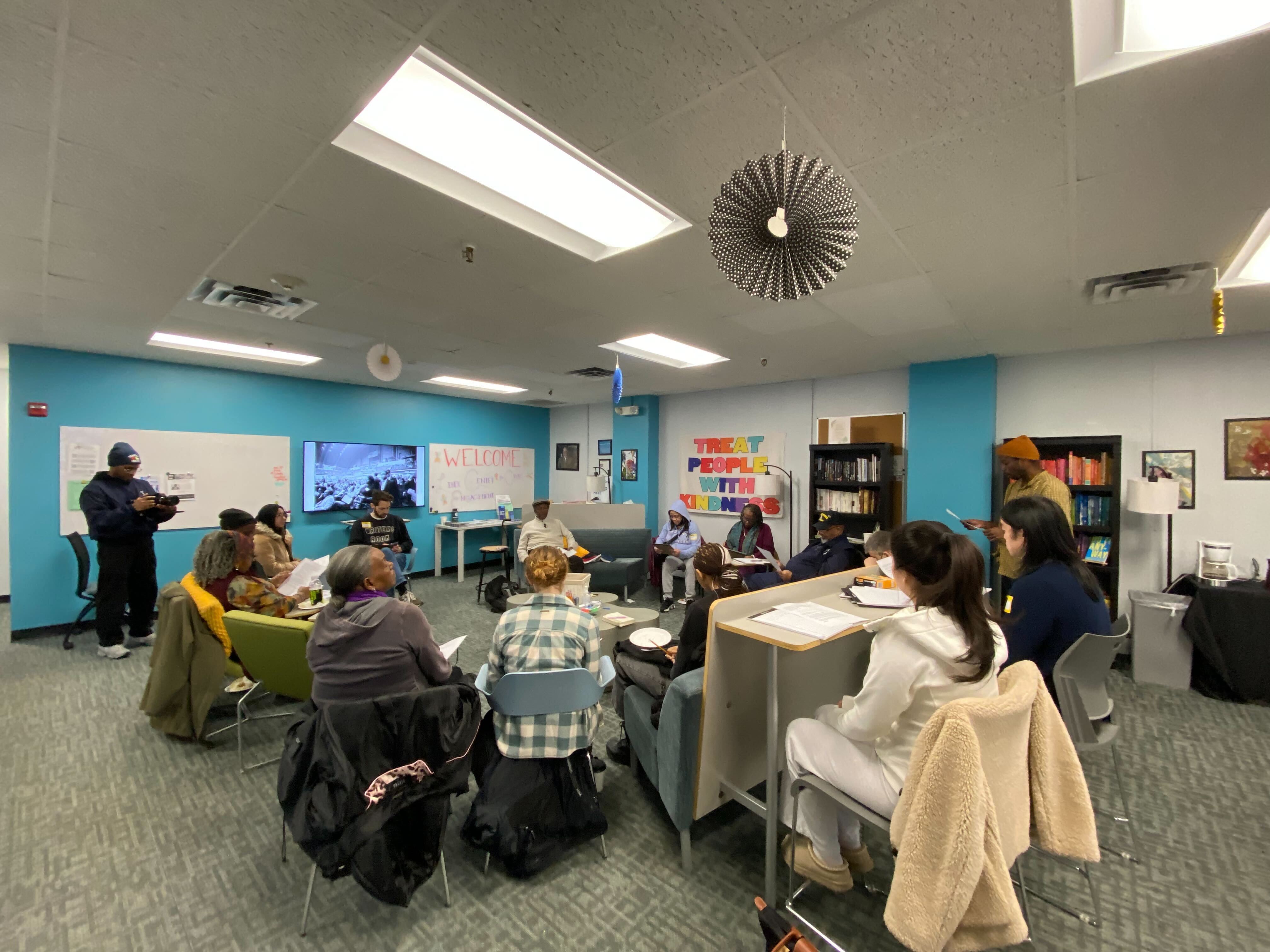 A group of people sit in a room listening to a speaker. The speaker has dark skin and is sitting on a couch. He is wearing a sweater and a hat. 