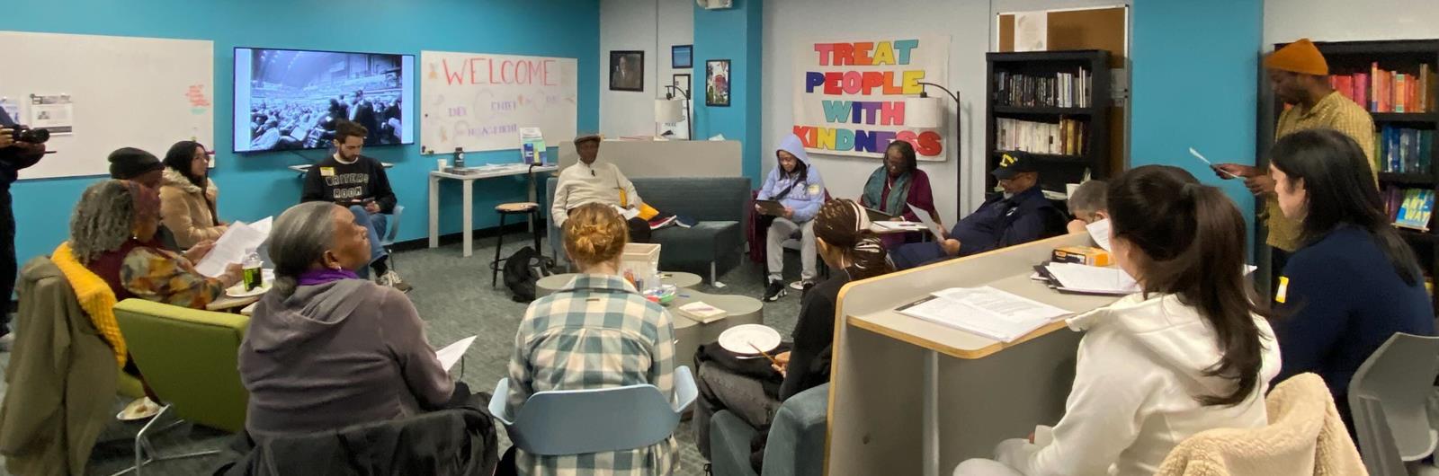 A group of people sit in a room listening to a speaker. The speaker has dark skin and is sitting on a couch. He is wearing a sweater and a hat. 