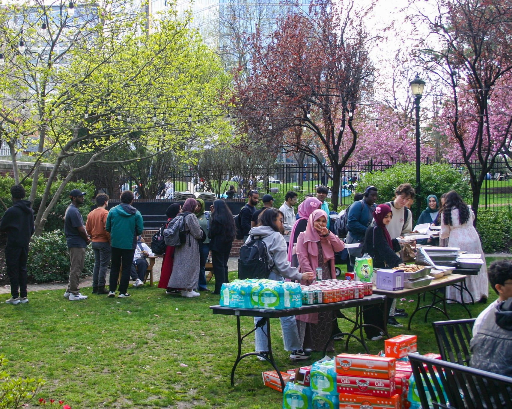 A large group of people are participating in a festival in an outdoor space with green grass and trees. In the forefront of the image is a table where drinks in plastic bottles are being handed out. 