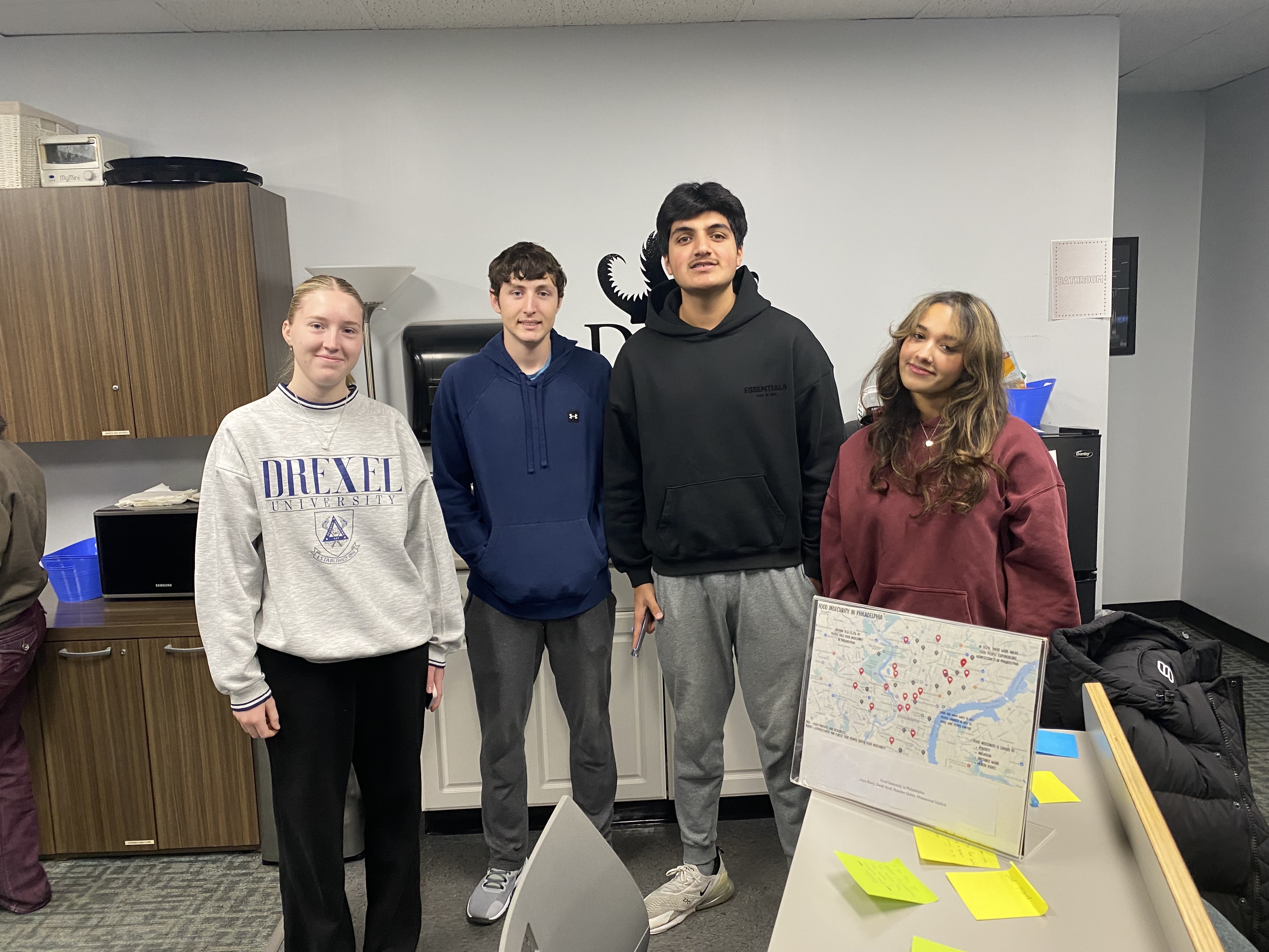 Four students standing together in an office space, smiling at the camera. One person wears a gray Drexel University sweatshirt, while the others wear hoodies and casual clothing. A table in the foreground holds sticky notes and a display board with a city map.