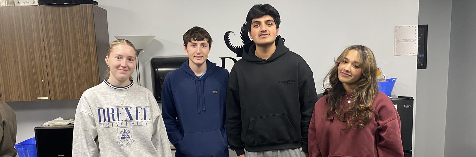 Four students standing together in an office space, smiling at the camera. One person wears a gray Drexel University sweatshirt, while the others wear hoodies and casual clothing. 