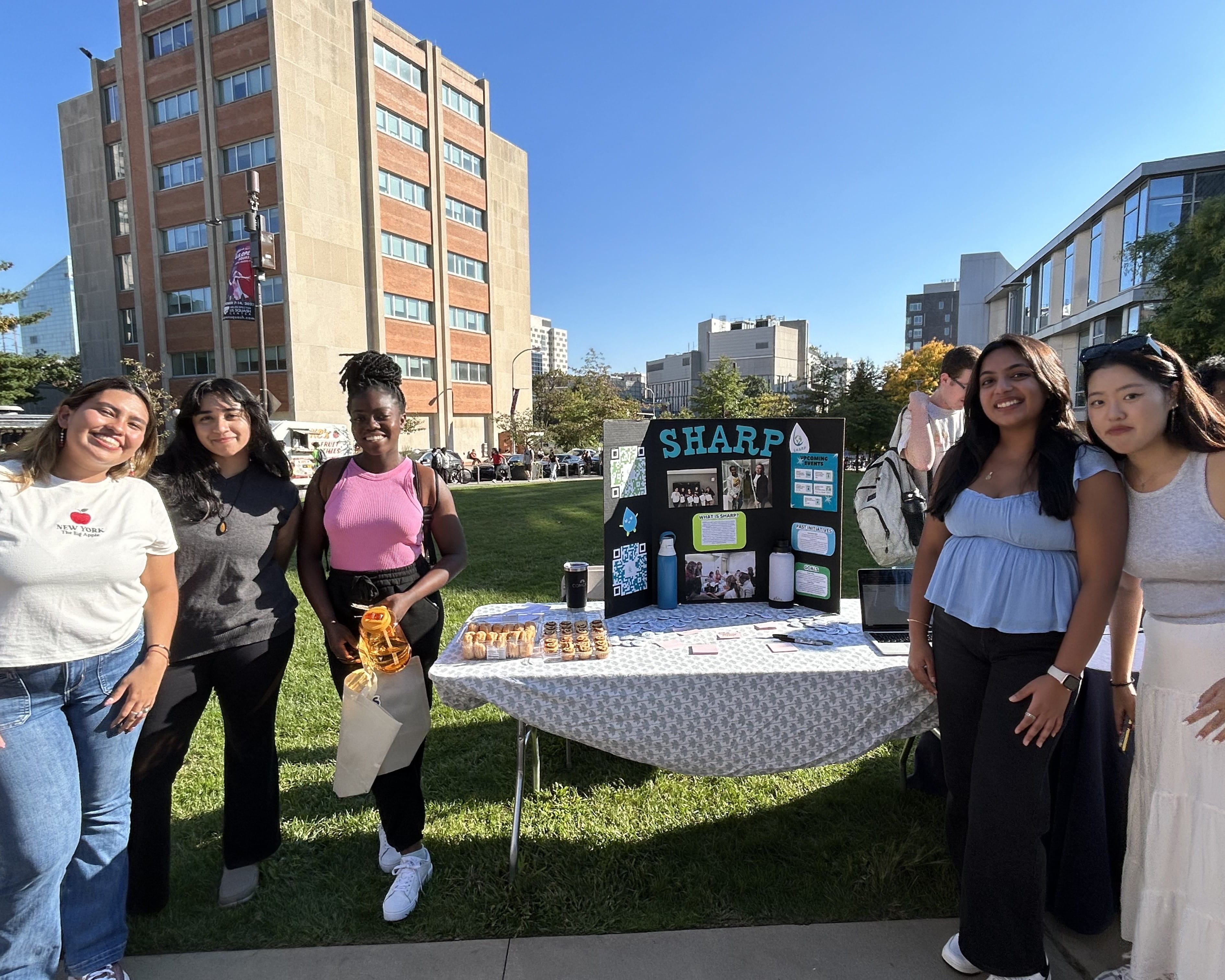 A group of 5 women are standing on either side of a table. The table has a white tablecloth and a display board collage that says "SHARP". The women are all in various different outfits and they are all smiling. 