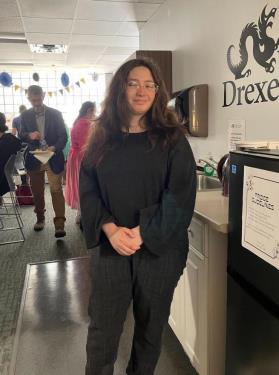 Izzy has long brown hair and round glasses and is smiling and clasping her hands together. In the background people are gathered in the Lindy Center for the End of Year Celebration. 