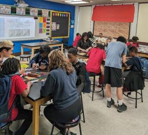 The image shows several tables in a classroom with people of various ages, skin colors, and clothing styles sitting and standing around the tables. It is an image of Drexel students from the Lindy Scholars program interacting with students at a local middle school. 