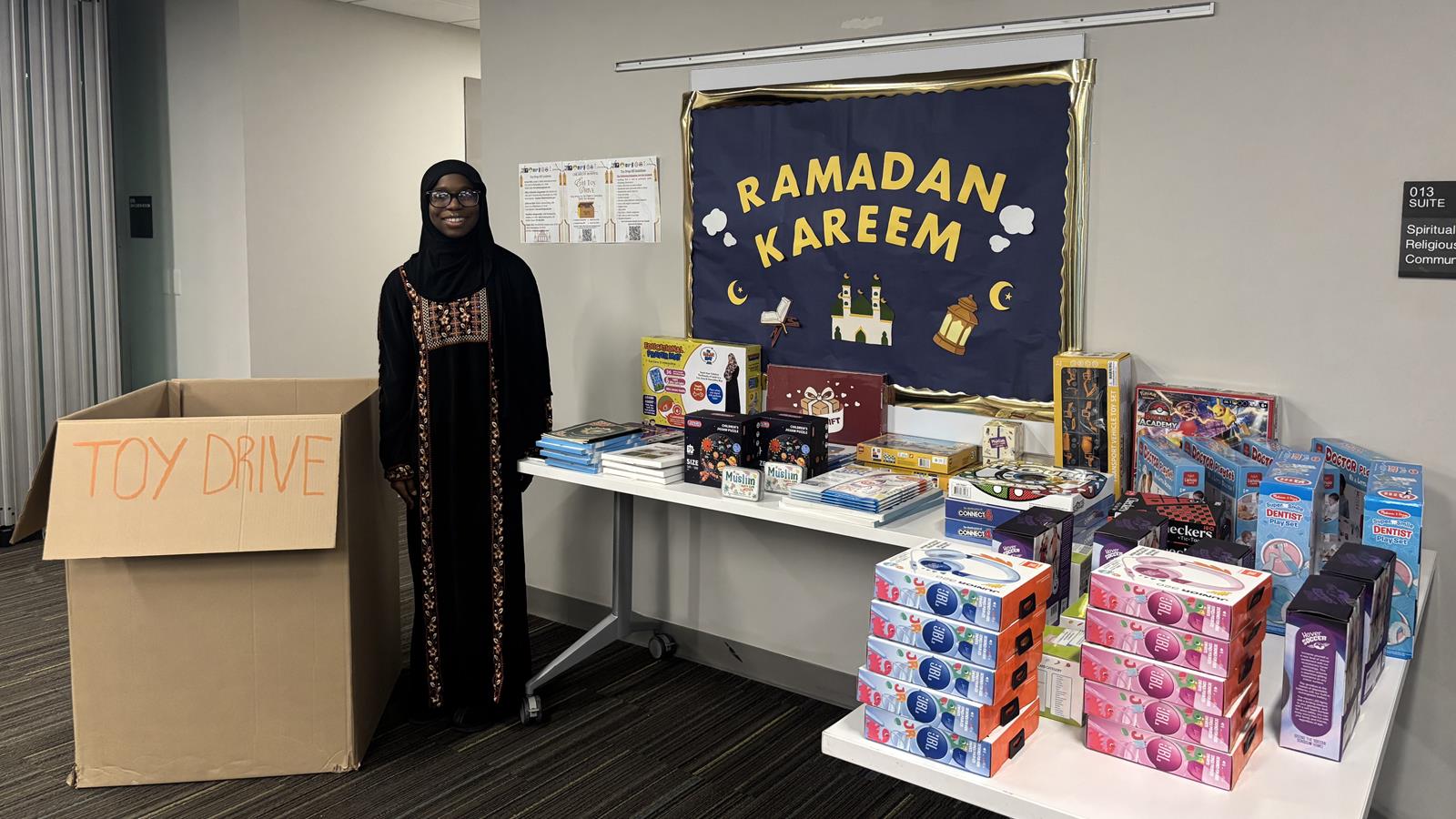 Taslim, a dark-skinned person in a black hijab and long black dress with glasses, stands smiling in front of a table that is covered in toys. There is a sign behind her saying "Ramadan Kareem" and a box next to her labeled "Toy Drive". 