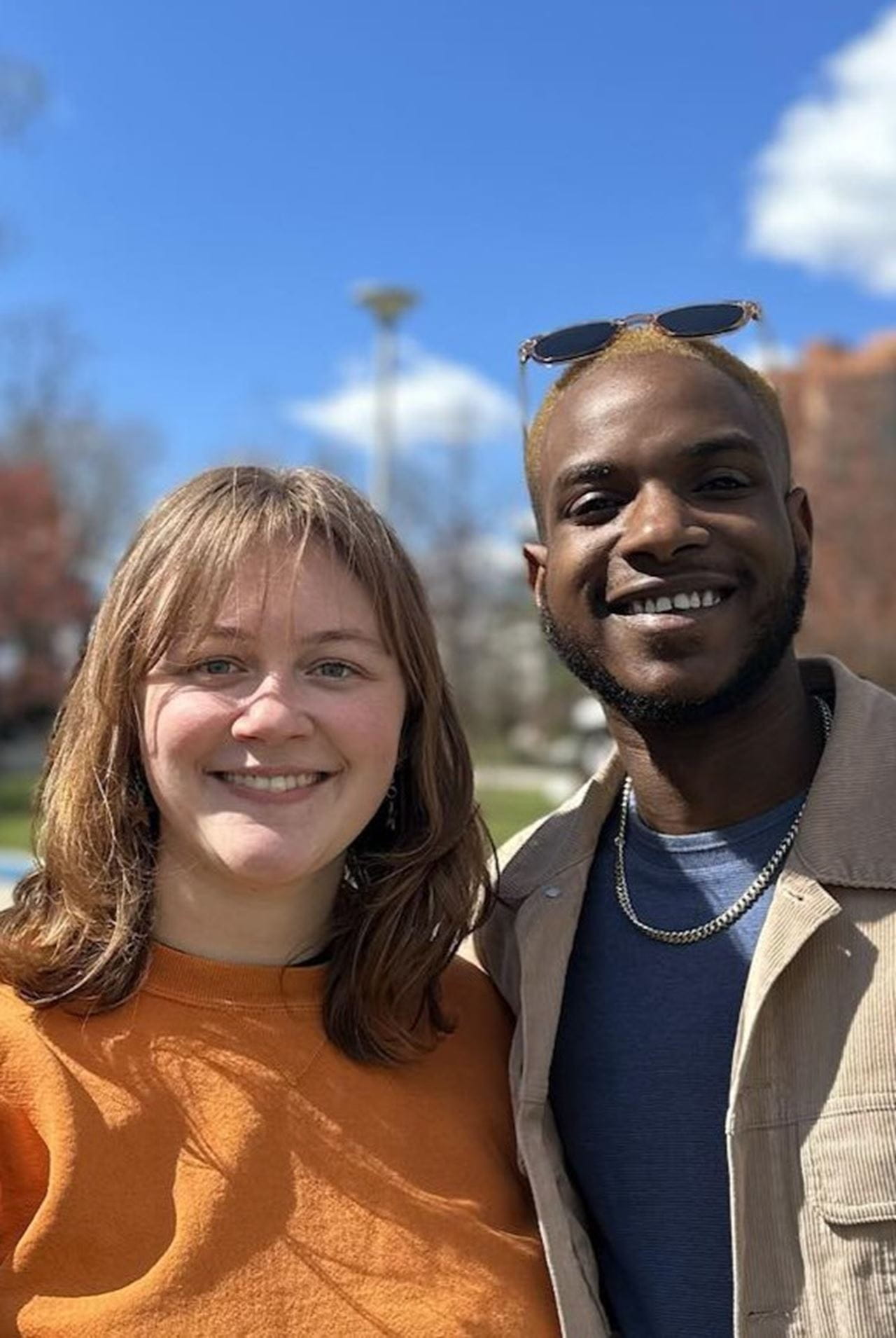 Two people stand together smiling. The person on the left is in an orange shirt and has white skin and light brown hair. The person on the right is in a blue shirt with a khaki blazer, has Black skin, and has sunglasses pushed up on their head. 