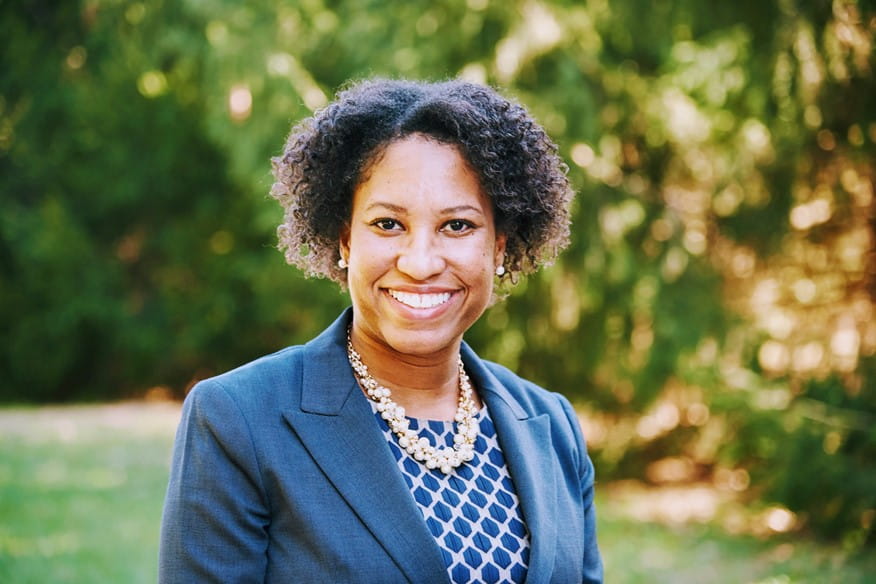 person standing outside smiling, wearing gray blazer, blue shirt, and necklace