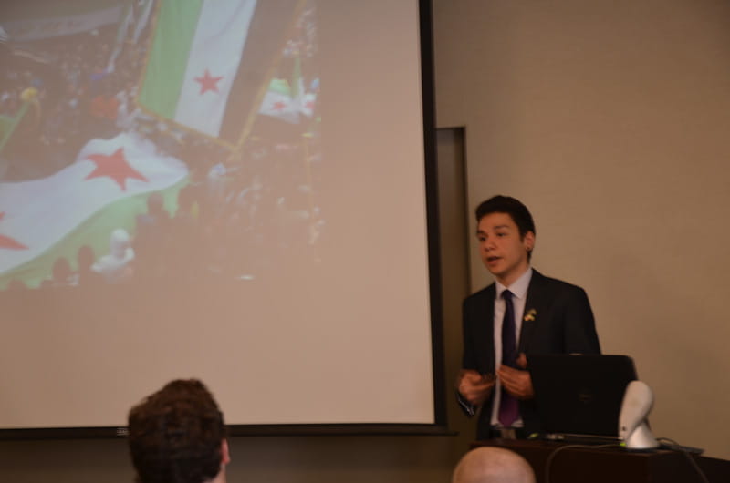 Hallak discussing demonstrations in Syria with a photo of one from his presentation flanking him.