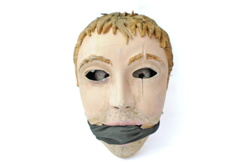 A mask depicting a face with a scar of an eye, with eyeballs deep within the eye holes and a gag over the mouth.