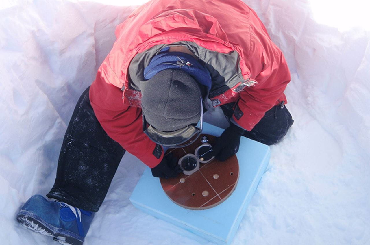 A man in winter gear leaning over a sensor in a howl in the snow and ice.