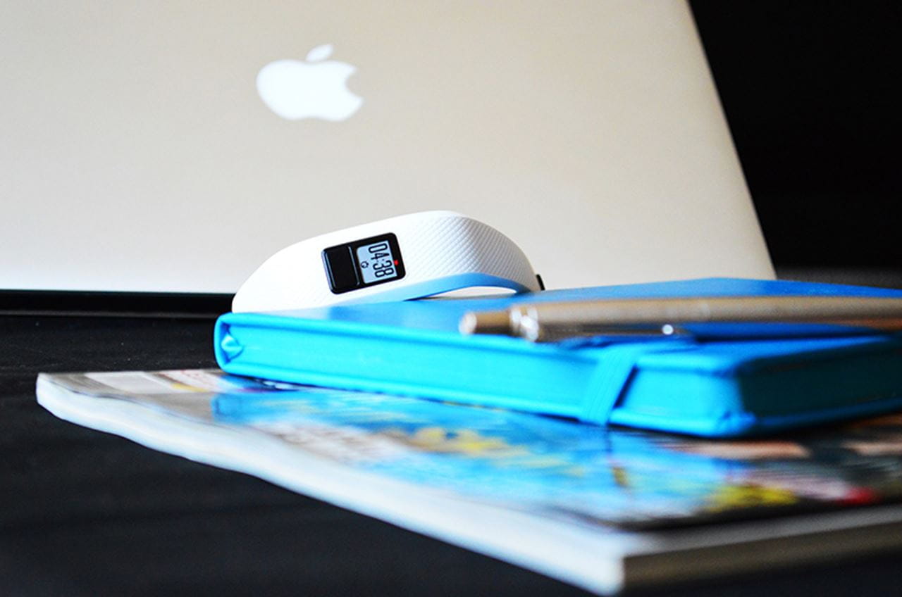 Wrist worn fitness tracker sitting atop a notebook, pen and magazine. All items are siting in front of a lap top. 