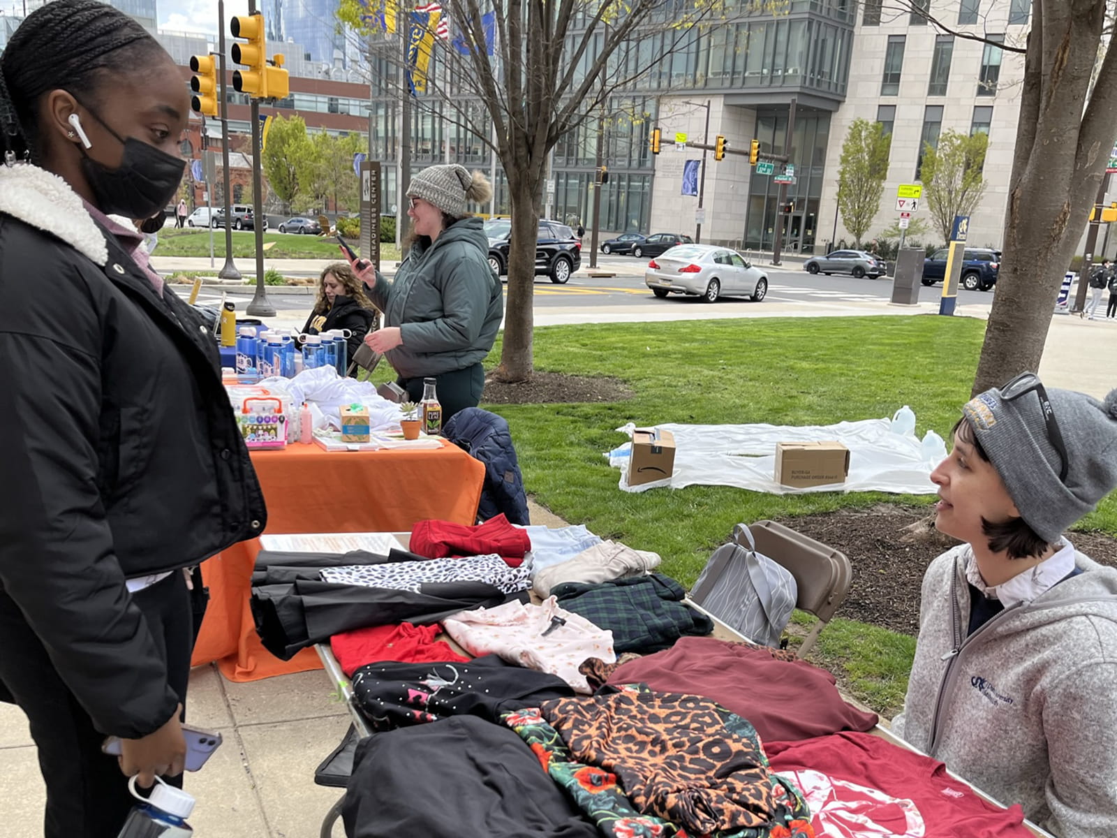 On Lancaster Walk, a person attending EarthFest speaks to someone seated at a table with clothing. 