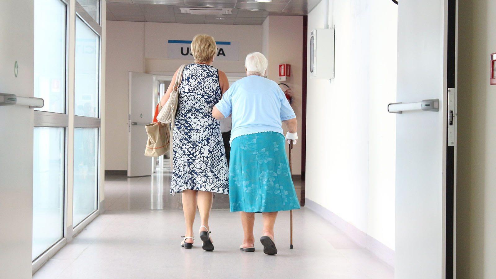 Older woman walking with cane down hospital hallway with younger woman