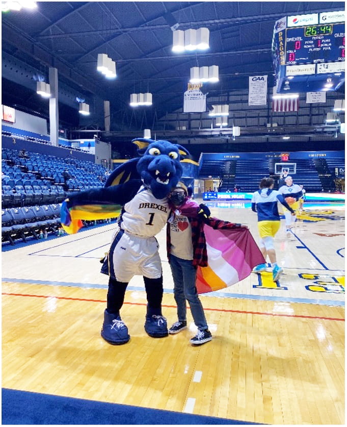 Masked student poses with Mario the Dragon, Drexel's mascot, on the basketball court in front of two female players.
