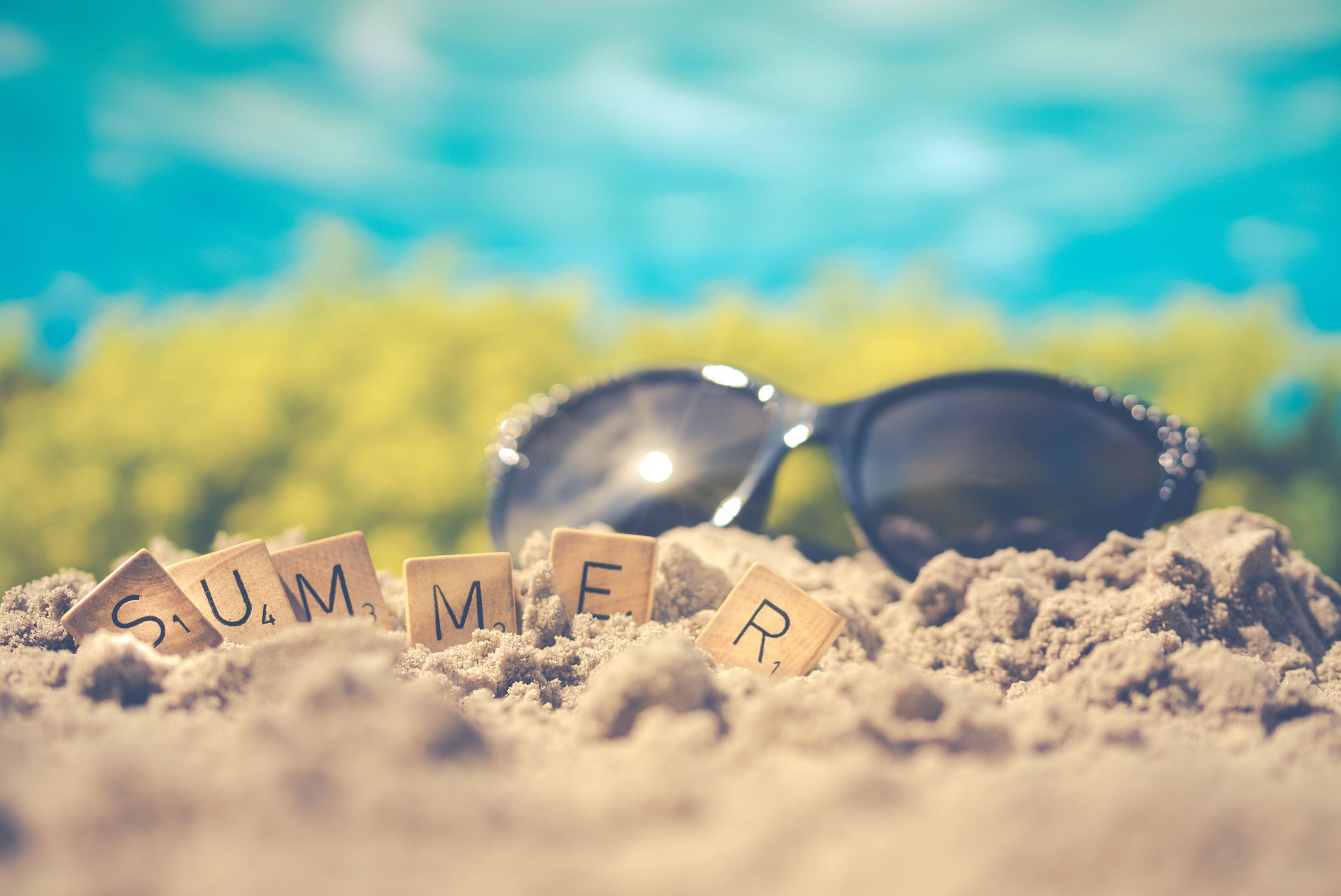 Scrabble blocks spelling out "summer" in the sand with sunglasses in the background.