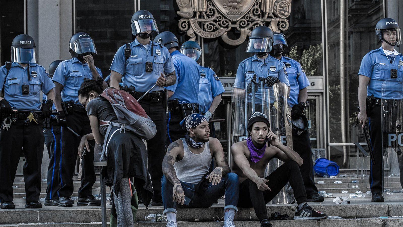 Two men sit in front of a crowd of police officers.