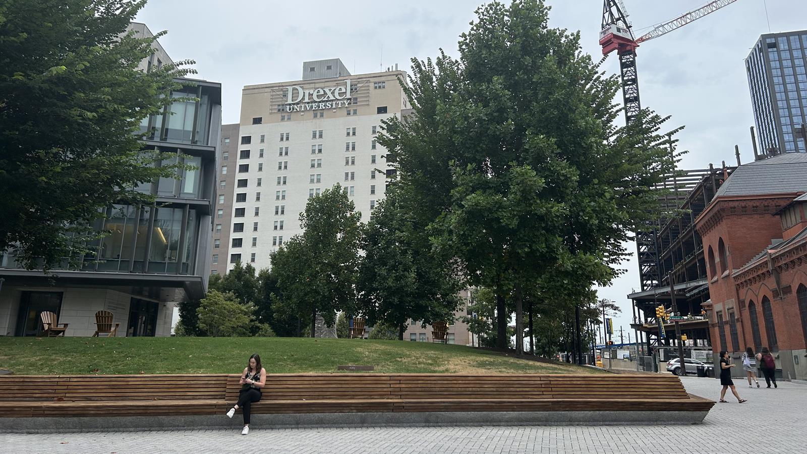 A woman sits on a brown bench in a paved outdoor area of Drexel University's Korman Quad with a construction crane in the background.