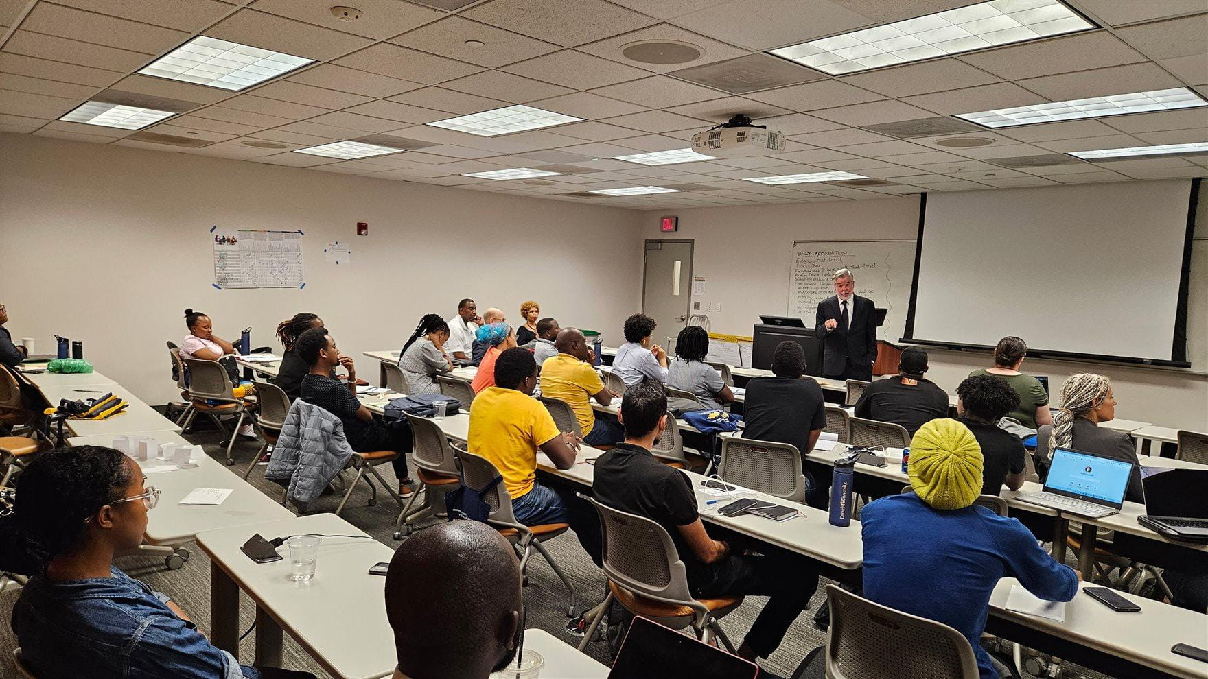 Four rows of people sit in long tables in a classroom as Drexel President John Fry stands at the front of the room addressing them. 