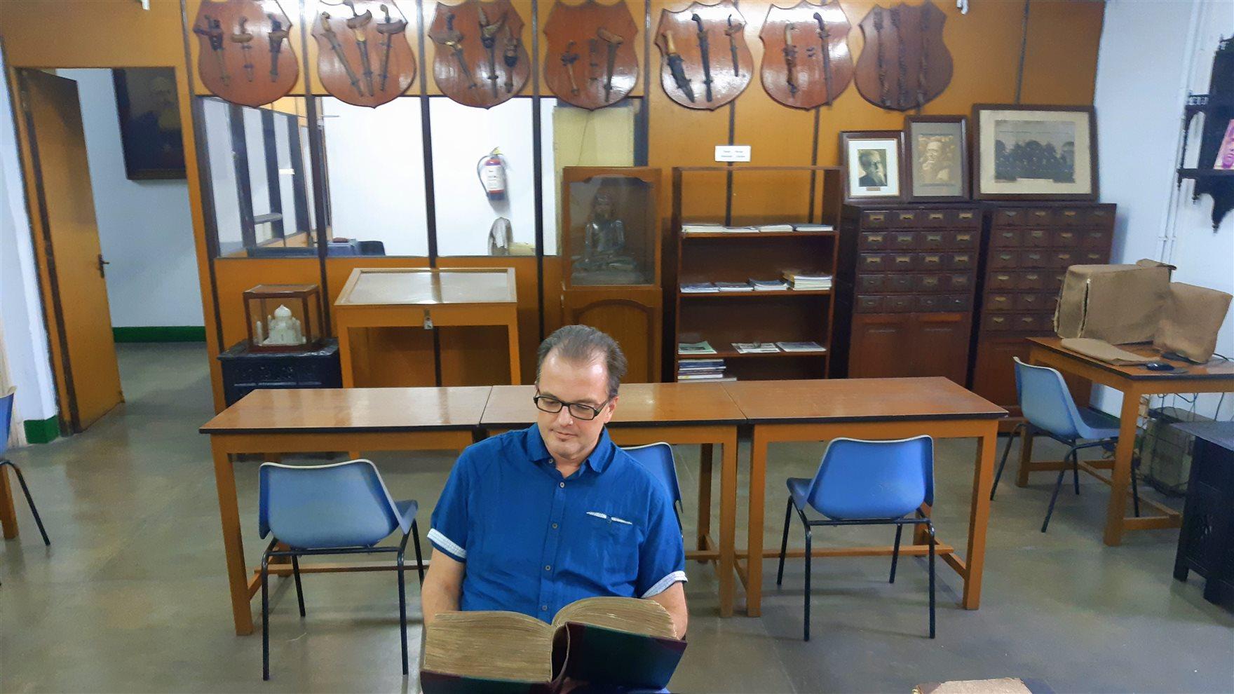A man in a blue shirt seated at a table looking at a large book in a library.