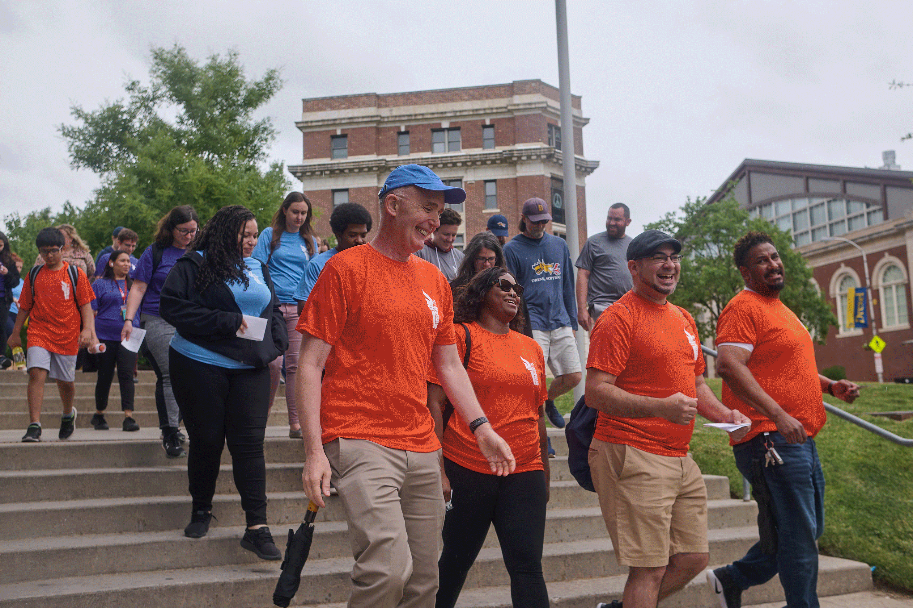 GIf of images of a group of people in orange shirts leading a group down stairs; people playing Baggo on a basketball court; two people playing table tennis; people in purple andornage shirts on a sandf volleyball court oplaying and a woman petting a dog.