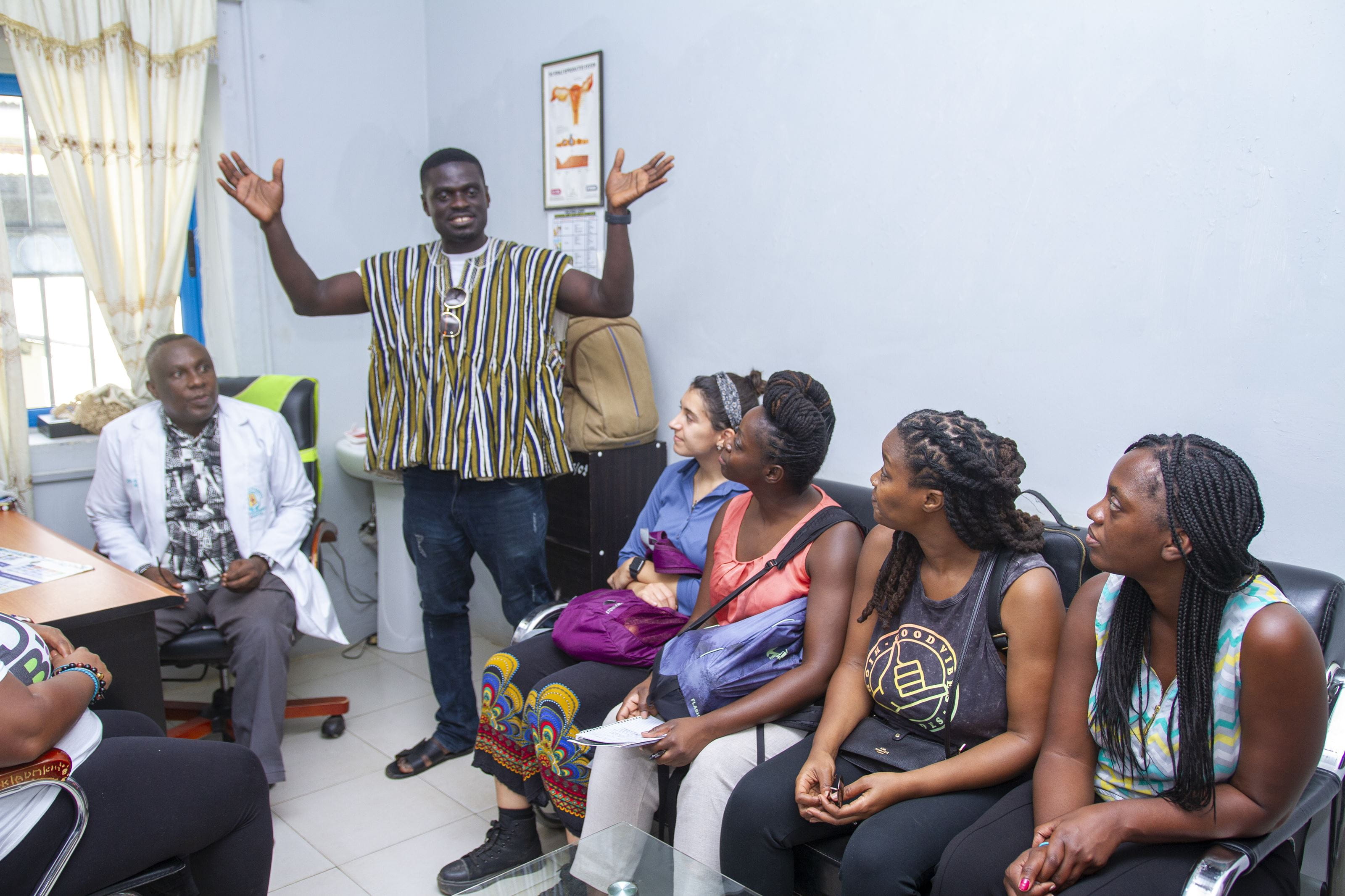 Four women sit in a sofa as a man raises his hand to the left of them and a man in a white coat sits and watches.