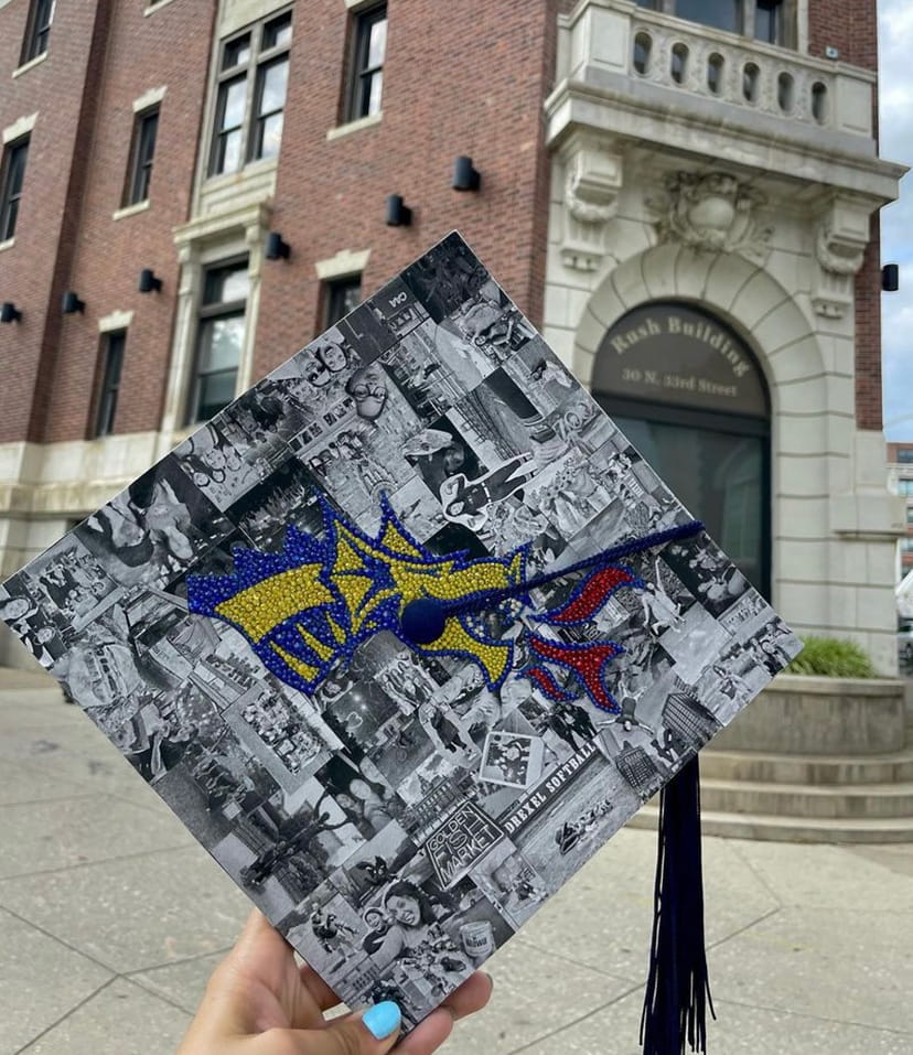 Someone holds a commencement cap in front of the Rush Building with the bedazzled Drexel Athletics logo over black and white photos. 